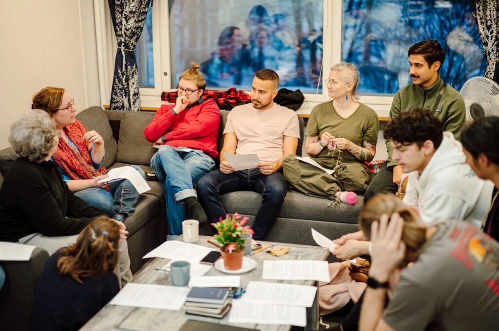 Kuvassa kymmenen ihmistä istuu ringissä, osa sohvalla, osa lattialla. Yksi heistä puhuu ja toiset kuuntelevat. Heidän kuvansa heijastuu taustalla olevasta isosta ikkunasta talvimaiseman päälle.