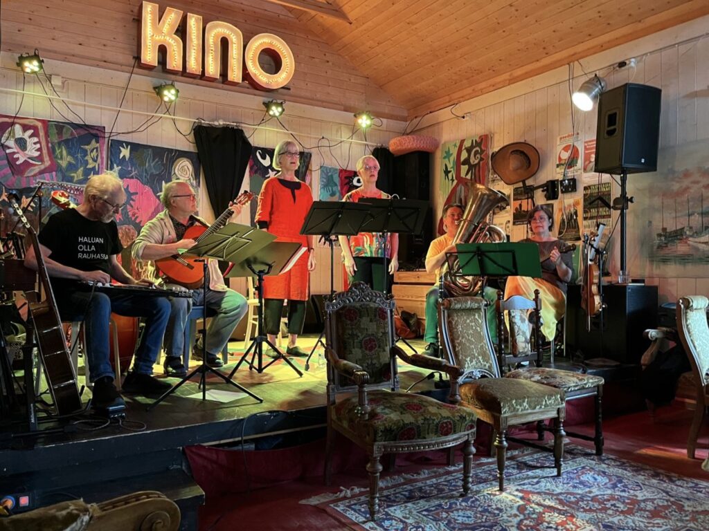 Kuvassa on kuusi ihmistä esiintymislavalla. Keskellä kaksi heistä laulaa, ja reunalla olevat soittavat eri instrumentteja. Takaseinällä on iso Kino-valo.