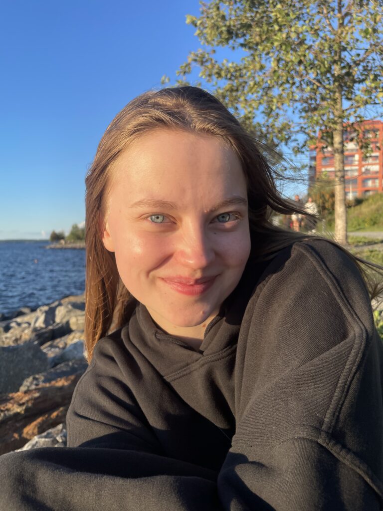 Lähikuvassa on yksi henkilö. Taustalla näkyy lehtipuu, kerrostalo, vettä ja sinistä taivasta.