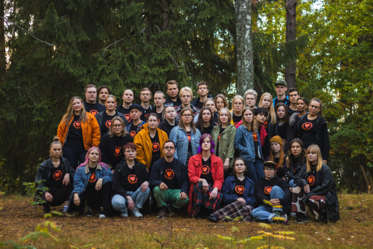 Kuvassa iso ryhmä nuoria ihmisiä on kokoontunut yhteen siten, että edessä olevat istuvat maassa ja takana olevat seisovat. Taustalla näkyy lehti- ja havupuita.