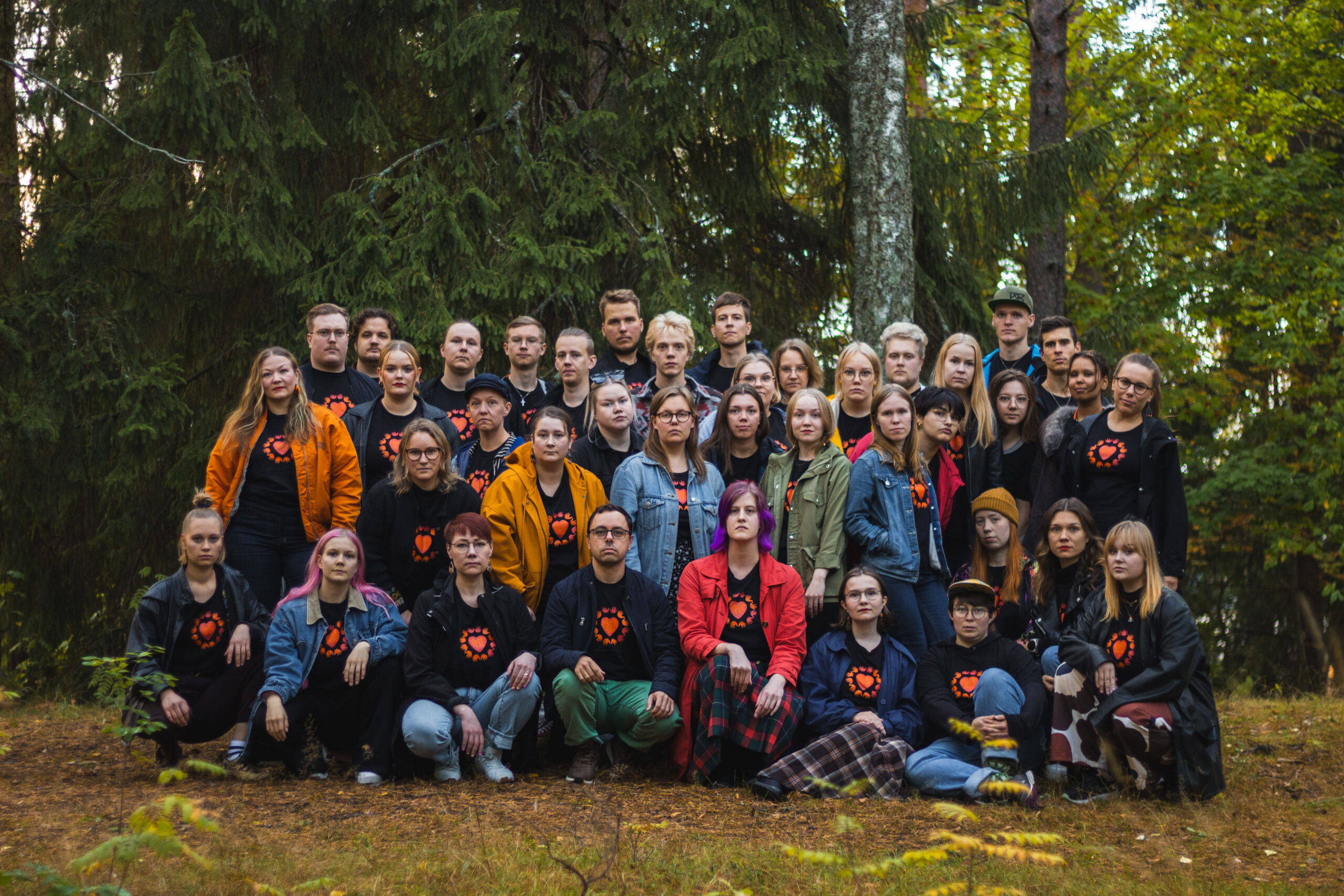 Kuvassa iso ryhmä nuoria ihmisiä on kokoontunut yhteen siten, että edessä olevat istuvat maassa ja takana olevat seisovat. Taustalla näkyy lehti- ja havupuita.