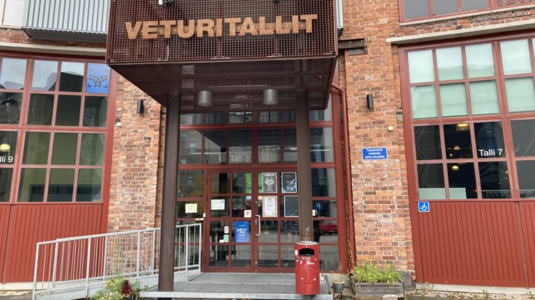 Kuvassa on punatiilisen rakennuksen sisäänkäynti, jonka yllä lukee teksti VETURITALLIT. Sekä ovessa että sen molemmin puolin on paljon lasiruutuja.
