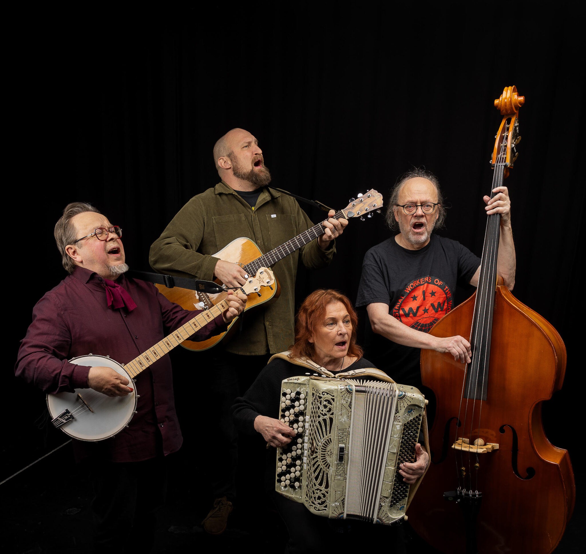 Kuvassa on neljä ihmistä, jotka laulavat ja soittavat eri instrumentteja. Yksi heistä istuu edessä ja soittaa harmonikkaa, ja hänen takanaan seisovat soittavat vasemmalta lukien banjoa, kitaraa ja kontrabassoa.